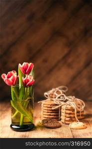 Simple rustic still life with blooming red tulips in a green glassand stacks various cookies on a dark wooden table. Fine art rustic background. Close Up.. Simple rustic still life of red tulips in a green glass and stacks various cookies on wooden table.