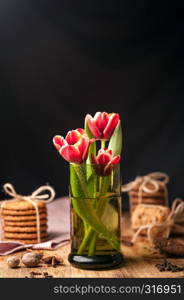 Simple rustic still life with blooming red tulips in a green glassand stacks various cookies on a dark wooden table. Fine art rustic background. Close Up.. Simple rustic still life of red tulips in a green glass and stacks various cookies on wooden table.
