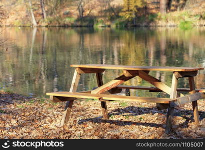Simple wooden table with picnic benches in the open air on a background of fallen oak leaves near a forest lake on a warm autumn day in blur with copy space.. Wooden table with picnic benches in the open air on the background of fallen oak leaves near a forest lake.
