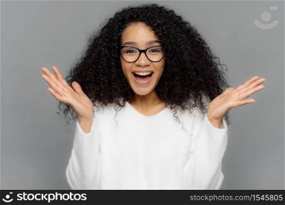 Sincere emotions and feelings concept. Optimistic lovely young female spreads hands, laughs happily at sees something pleasant, has curly bushy hairstyle, wears spectacles and white casual jumper
