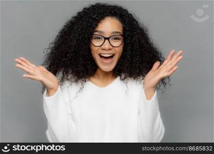 Sincere emotions and feelings concept. Optimistic lovely young female spreads hands, laughs happily at sees something pleasant, has curly bushy hairstyle, wears spectacles and white casual jumper
