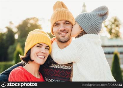 Sincere emotions. Little cute girl in knitted hat and white warm sweater kisses her father with love. Friendly affectionate couple pose together outdoors, smile happily, have wonderful relationships