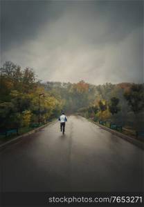 Single man running alone on an alley in the autumn park. Practicing sport outdoors in a rainy and misty day. Gloomy fall season mood. Healthy lifestyle workout jogging activity.