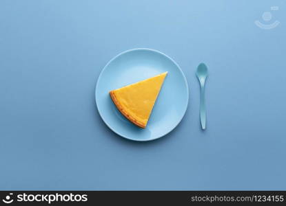 Single piece of cheesecake on a blue plate on blue background. Above view of dessert plate with cheesecake slice and blue spoon. Famous cheese cake.