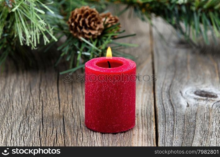 Single red candle with flame and evergreen setting on rustic wood. Selective focus on front part of candle and flame.