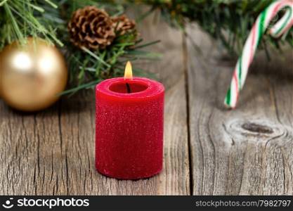 Single red candle with flame and ornaments in evergreen setting on rustic wood. Selective focus on front part of candle and flame.