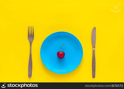 Single ripe cherry on blue plate, knife, fork. Still life on yellow background, top view, Flat lay Copy space Concept summer and diet, losing weight