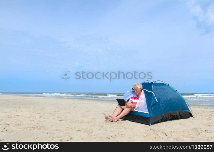 Single senior man with digital tablet camping in shelter at the beach