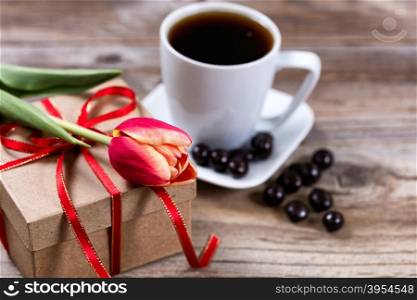 Single tulip resting on gift box with a cup of coffee and dark chocolate in background on rustic wood. Selective focus on front part of flower.