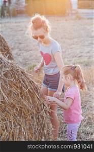 Sisters, teenage girl and her younger sister playing together with hay bale outdoors in the field in the countryside. Candid people, real moments, authentic situations