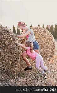 Sisters, teenage girl and her younger sister pushing hay bale playing together outdoors in the countryside. Candid people, real moments, authentic situations