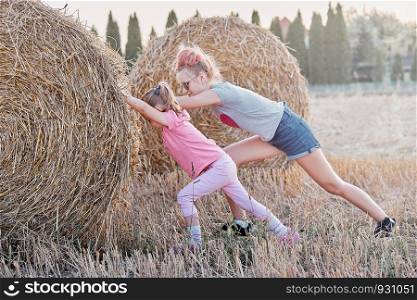 Sisters, teenage girl and her younger sister pushing hay bale playing together outdoors in the countryside. Candid people, real moments, authentic situations
