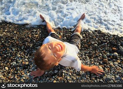 sitting teenager boy looking upwards on stone seacoast, wets feet in water, sitting by back