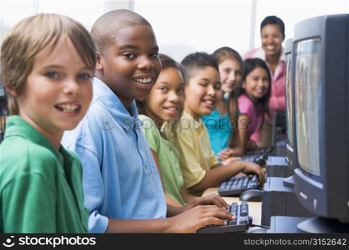 Six children at computer terminals with teacher in background (selective focus/high key)