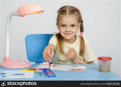 Six year old girl Europeans draws paints on a sheet of sitting at the table. Six year old girl dunks brush in the paint