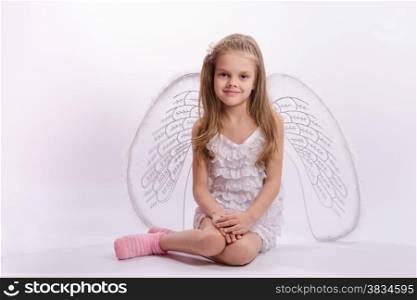 Six year old girl in a bright angel costume with wings on a white background. Sitting girl in an angel costume on a white background