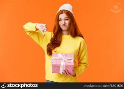 Skeptical and awkward cute redhead picky girl dont like christmas gift from aunt, show thumb-down, grimacing with uncomfortable judgemental expression, holding pink ugly present, orange background.. Skeptical and awkward cute redhead picky girl dont like christmas gift from aunt, show thumb-down, grimacing with uncomfortable judgemental expression, holding pink ugly present, orange background