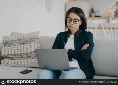 Skeptical businesswoman working on laptop reading email, crossed arms sitting on sofa. Pensive female freelancer in glasses reading bad news, solving working problem looking at computer screen at home. Skeptical businesswoman working on laptop reading email, crossed arms, sitting on couch at home