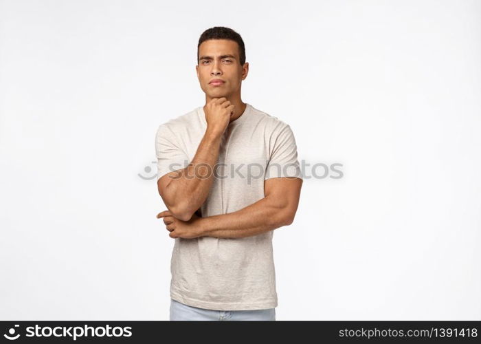 Skeptical, suspicious handsome strong man in t-shirt, have doubts, hold arm under chin, squinting suspect something wrong, have hesitations or concernes, standing judgemental white background.. Skeptical, suspicious handsome strong man in t-shirt, have doubts, hold arm under chin, squinting suspect something wrong, have hesitations or concernes, standing judgemental white background