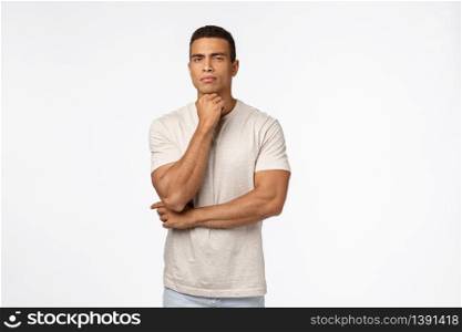 Skeptical, suspicious handsome strong man in t-shirt, have doubts, hold arm under chin, squinting suspect something wrong, have hesitations or concernes, standing judgemental white background.. Skeptical, suspicious handsome strong man in t-shirt, have doubts, hold arm under chin, squinting suspect something wrong, have hesitations or concernes, standing judgemental white background