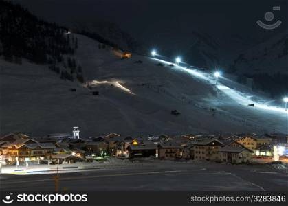 Ski village at night with slope lights, cross-country ski run, buildings, ice monument - shot in Livigno, Italian Alps