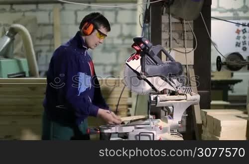 Skilled carpenter in protective workwear measuring wooden plank with ruler and pencil for cutting. Master joiner sawing board with circular saw machine in woodworking shop.