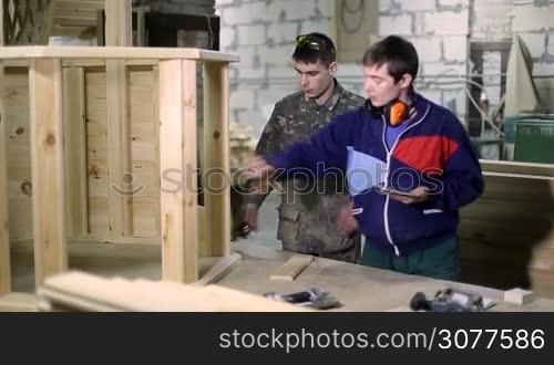 Skilled carpenter using digital touchpad and giving instructions to his coworker at carpentry shop. Joiner measuring product height with tape while his senior colleague working on tablet pc, explaining project details.