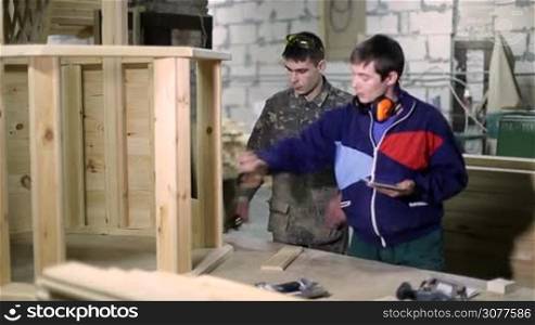 Skilled carpenter using digital touchpad and giving instructions to his coworker at carpentry shop. Joiner measuring product height with tape while his senior colleague working on tablet pc, explaining project details.