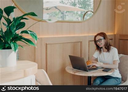 Skilled female freelancer has video conversation on laptop computer in cafeteria enjoys friendly web calling uses earphones poses at table with coffee and croissant connected to public internet. Skilled female freelancer has video conversation on laptop computer