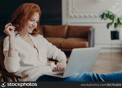 Skilled positive female student creats article for homework, sits in coworking space, has pleased expression, works on laptop computer, pose over home interior, prepares for writing course work