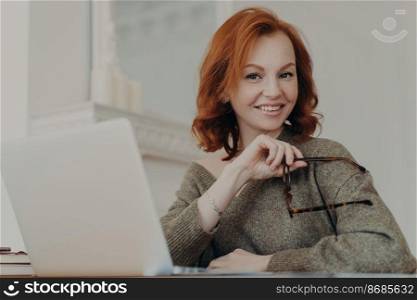 Skilled successful redhead woman holds eyewear for vision protection, sits in front of opened laptop computer, poses at coworking space, wears sweater, uses modern device. Distance work concept