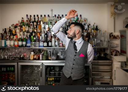 Skilled young bearded male bartender in stylish uniform performing trick with bottle of alcoholic drink while preparing cocktail at bar counter. Barman doing trick with bottle