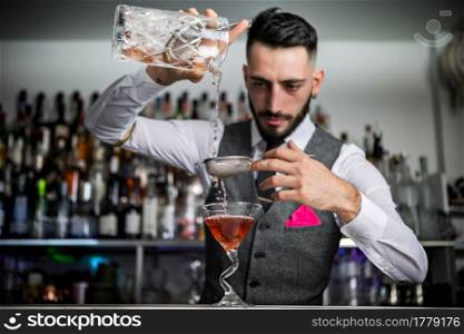 Skilled young Hispanic barman in elegant clothes filling goblet with alcohol cocktail through metal sieve while working at bar counter. Bartender with sieve pouring drink into glass