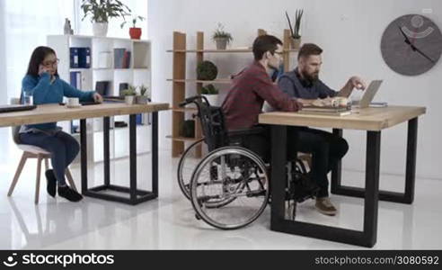 Skillful disabled man in wheelchair discussing new startup project on laptop pc with coworker while asian female colleague talking on phone in creative office. Handicapped freelancer working in office