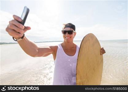 skimboarding, water sport and people concept - happy smiling young man with skimboard and smartphone taking selfie on summer beach. happy young man with skimboard on summer beach
