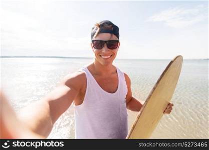 skimboarding, water sport and people concept - happy smiling young man with skimboard taking selfie on summer beach. happy young man with skimboard on summer beach
