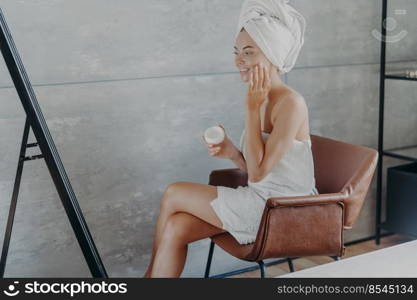 Skin care concept. Indoor shot of pretty smiling woman sits in comfortable armchair opposite mirror, applies cosmetic cream on face, wears wrapped towel on head, wants to have well groomed complexion