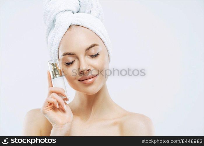 Skin care, cosmetics concept. Photo of relaxed healthy young European woman stands with closed eyes, holds bottle of cosmetic product for elastic skin, poses half naked against white background.
