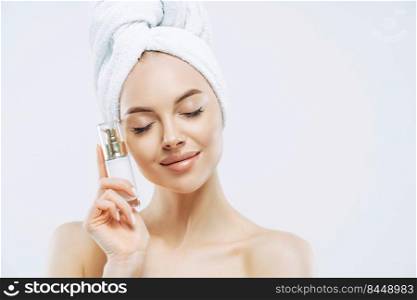 Skin care, cosmetics concept. Photo of relaxed healthy young European woman stands with closed eyes, holds bottle of cosmetic product for elastic skin, poses half naked against white background.