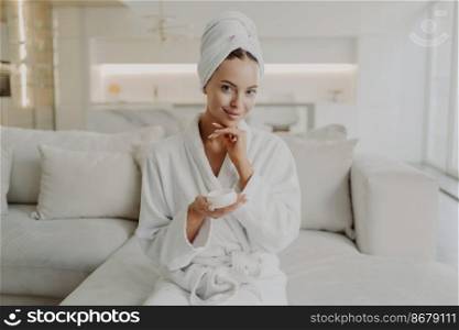 Skincare and beauty concept. Portrait of young attractive woman in bathrobe and towel on head holding cream jar and smiling at camera while sitting on sofa in living room doing cosmetic procedures. Young happy woman holding cosmetic face cream and smiling at camera while relaxing on sofa at home