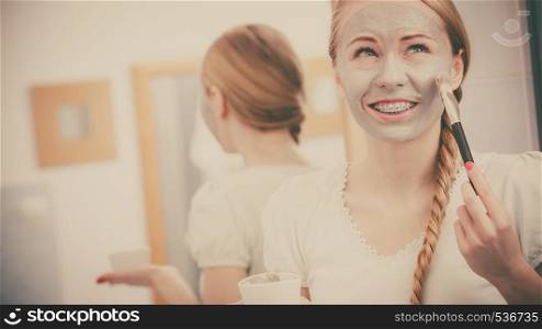 Skincare. Blonde woman in bathroom applying with brush gray clay mud mask to her face. Young lady taking care of skin. Spa beauty wellness.. Woman applying with brush clay mud mask to her face