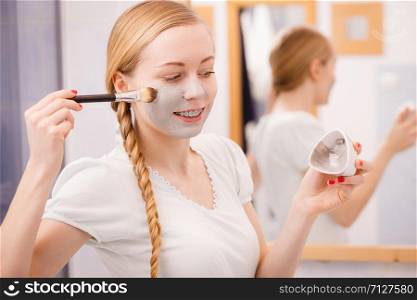 Skincare. Blonde woman in bathroom applying with brush gray clay mud mask to her face. Young lady taking care of skin. Spa beauty wellness.. Woman applying with brush clay mud mask to her face