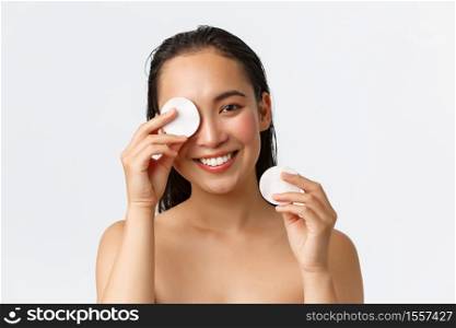 Skincare, women beauty, hygiene and personal care concept. Close-up of attractive naked asian woman in bathroom, wipe-off makeup with cotton pads and smiling camera, white background.. Skincare, women beauty, hygiene and personal care concept. Close-up of attractive naked asian woman in bathroom, wipe-off makeup with cotton pads and smiling camera, white background