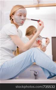 Skincare. Young woman wearing nightwear sitting on sink in bathroom, applying with brush gray clay mud mask to her face. Teen girl taking care of skin. Spa beauty wellness.. Woman applying with brush clay mud mask to her face
