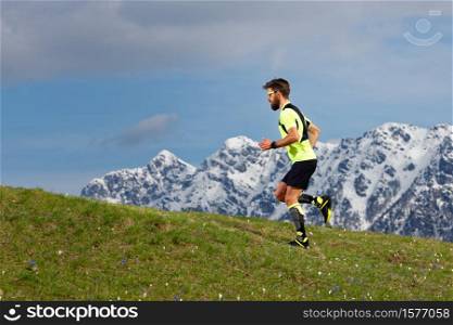 Skyrunning a bearded man in a spring meadow with snow mountains background.