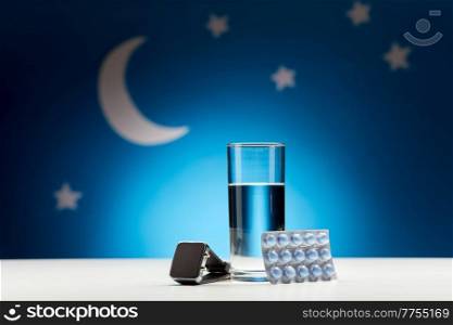 sleep disorder, bedtime and medicine concept - close up of glass of water, sleeping pills and smart watch over moon and night stars on blue background. water, sleeping pills and smart watch at night