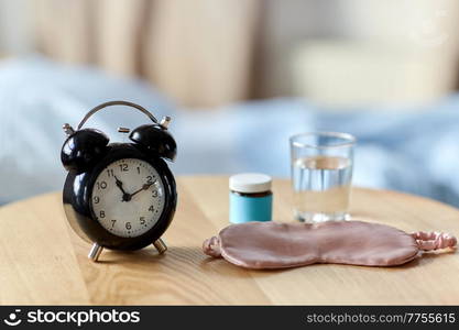 sleep disorder, bedtime and morning concept - close up of alarm clock, eye sleeping mask, glass of water and soporific medicine on night table at home. alarm clock, mask and sleeping pills on table