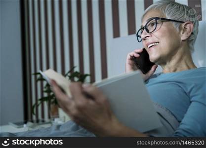 Sleep. Mature woman in bedroom, suffering from Insomnia, lying in bed, talking over the phone and reading a book. Sleep. Sleepless Mature Woman Suffering from Insomnia, Talking over the Phone and Reading a Book in Bed