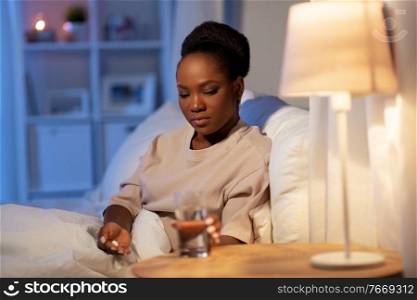 sleeping, health and people concept - african american woman with medicine and water at home at night. woman with medicine and water in bed at night