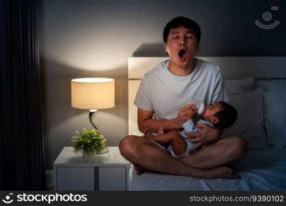 sleepy father yawning and feeding milk bottle to newborn baby on a bed at night